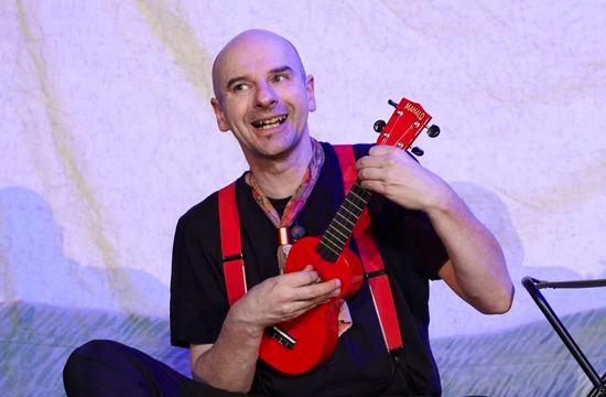 Eine Person mit roter kleiner Gitarre in der Hand sitzt auf dem Boden.